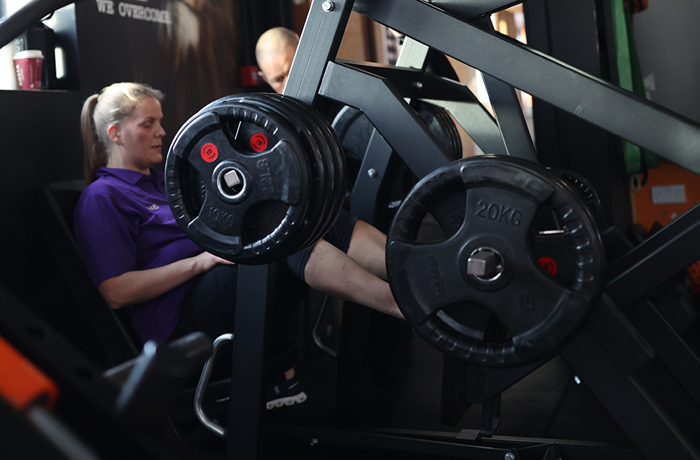A person is using a leg press machine in a gym. They are pushing weights labeled 20kg with their legs. The person is seated and wearing a purple shirt. Another individual in the background is slightly visible, seemingly observing or assisting.