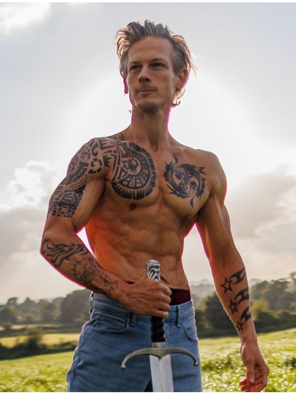 A shirtless man with tattoos, wearing jeans, stands in an open field holding a sword. The sun is shining behind him, casting a glow. Trees are visible in the background, and the sky is partly cloudy.